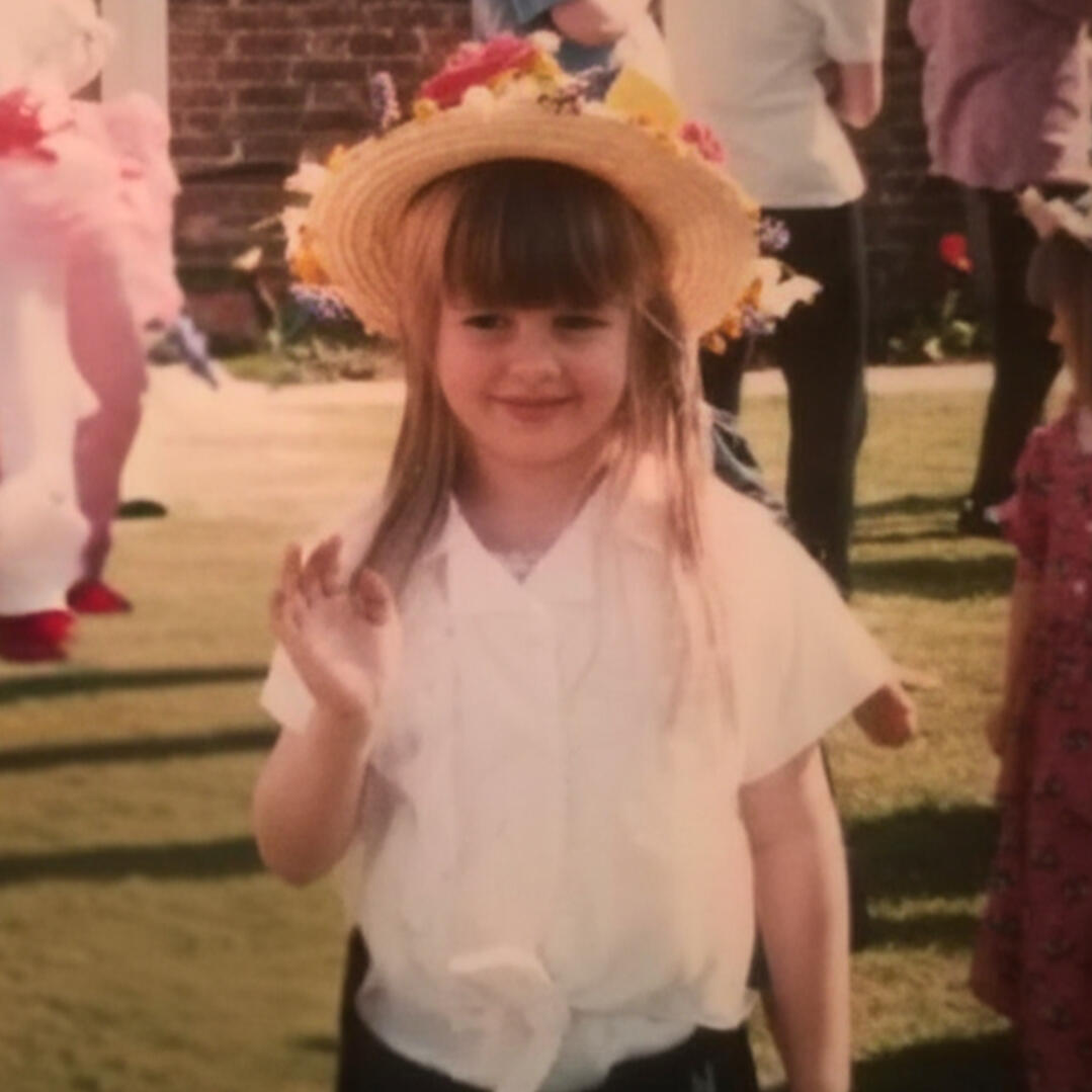 Picture of Katie as a small child, wearing a flowery sunhat and waving to the camera without making eye contact.